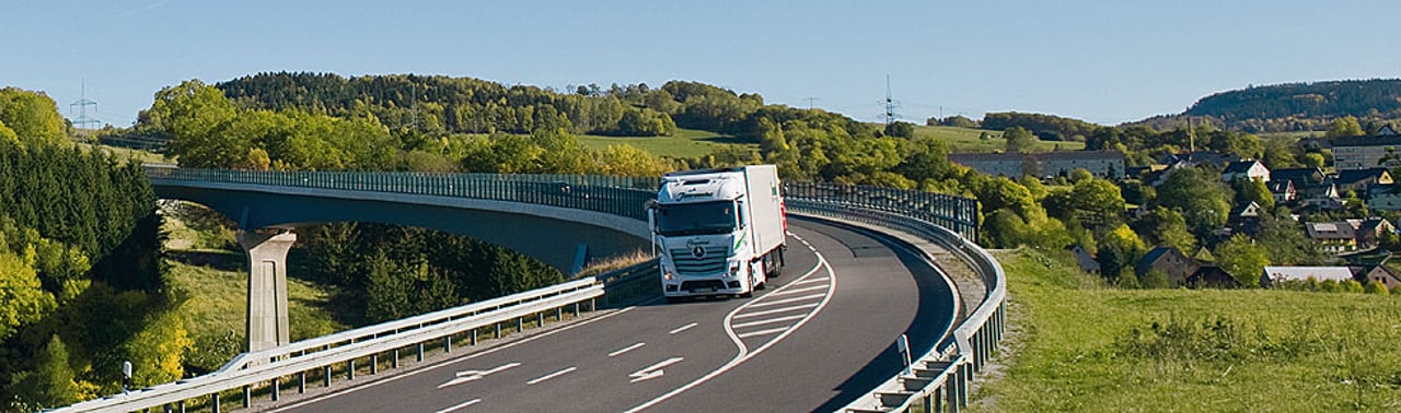 Ein weißer Lastwagen fährt auf einer geschwungenen Autobahnbrücke über ein bewaldetes Tal.