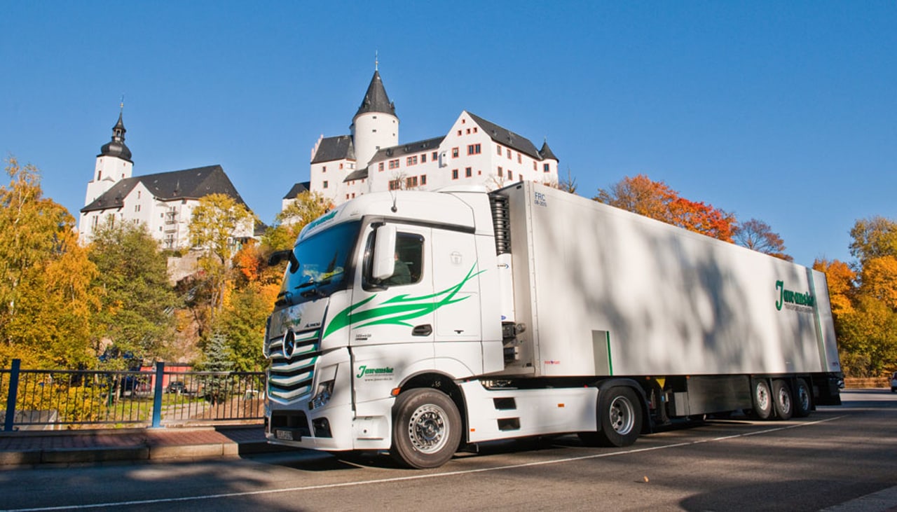 Weißer LKW mit grünen Streifen fährt vor einem weißen Schloss mit Türmen und bunten Herbstbäumen.